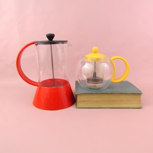May include: A red and clear French press coffee maker with a black lid and handle. Beside it is a clear glass teapot with a yellow lid and handle, resting on a vintage book. The background is a solid pink.