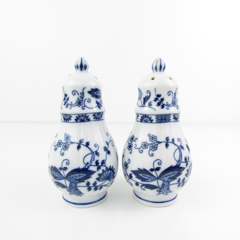 May include: A set of two white ceramic salt and pepper shakers with blue floral designs. The shakers have a bulbous shape with a small opening at the top for pouring.