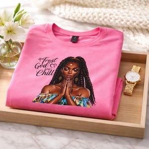 May include: A pink t-shirt with the words "Trust God and Chill" and an illustration of a woman with long braids. The shirt is folded on a wooden tray, next to a gold watch and a vase of white flowers.