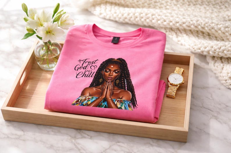 May include: A pink t-shirt with the words "Trust God and Chill" and an illustration of a woman with long braids. The shirt is folded on a wooden tray, next to a gold watch and a vase of white flowers.