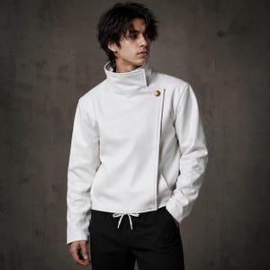 May include: A white jacket with a high collar and an asymmetrical front. It has a single gold button and a drawstring waist. The long-sleeved jacket is made of a smooth material. The model is wearing black trousers. The jacket is a stylish piece of outerwear.