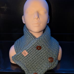 Peut inclure: Un col tricoté bleu-vert avec trois boutons décoratifs en bois. Le col est présenté sur une tête de mannequin. Une petite étiquette avec du texte est attachée au col. Les boutons ont un centre turquoise.