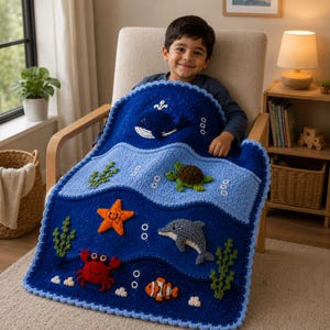 Op de afbeelding: Een rechthoekige gehaakte deken met een oceaanthema met donkerblauwe en lichtblauwe golvende secties. De geappliqueerde zeedieren omvatten een donkerblauwe walvis, een groene schildpad, een oranje zeester, een grijze dolfijn, een rode krab en een oranje anemoonvis.
