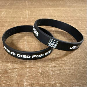 Könnte beinhalten: Zwei schwarze Armbänder aus Gummi mit weißem Text. Eines lautet "JESUS STARB FÜR MICH", das andere sagt "JESUS" und "IHM", mit einer kleinen quadratischen Grafik. Die Bänder sind kreisförmig und liegen auf einer Holzoberfläche.