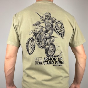 Könnte beinhalten: Khakifarbenes T-Shirt mit einer schwarzen Grafik eines Ritters auf einem Motorrad, der ein Schwert und einen Schild hält. Der Schild zeigt einen Löwen und das Wort "FAITH". Der Text "ARMOR UP. STAND FIRM." ist darunter gedruckt, mit Bezug auf Epheser 6.