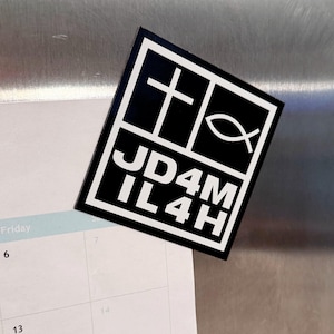 Könnte beinhalten: Schwarz-weißer rechteckiger Magnet mit einem Kreuz, einem Fischsymbol und dem Text "JD4M IL4H". Der Magnet wird auf einer Kalenderseite angezeigt.