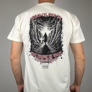 Könnte beinhalten: Cremefarbenes T-Shirt mit einem grafischen Design auf dem Rücken. Das Design zeigt eine Person, die auf ein Kreuz in einem dunklen Tal zugeht, mit dem Text "FEAR NO EVIL WHEN YOU WALK THROUGH THE VALLEY OF THE SHADOW OF DEATH".