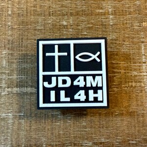 Könnte beinhalten: Ein quadratisches, schwarz-weißes Emblem mit einem Kreuz und einem Fischsymbol. Der Text "JD4M IL4H" wird in weißen Blockbuchstaben angezeigt. Das Emblem befindet sich auf einem Holzgrund.