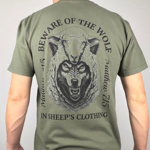 Könnte beinhalten: Olivgrünes T-Shirt mit einer detaillierten Grafik eines Wolfs im Schafspelz. Der Text "BEWARE OF THE WOLF" wölbt sich über dem Bild, mit "Matthew 7:15" und "IN SHEEP'S CLOTHING" ebenfalls auf dem Shirt gedruckt.