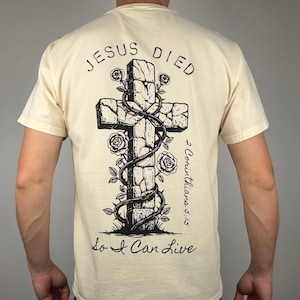 Könnte beinhalten: Cremefarbenes T-Shirt mit einer Grafik eines Steinkreuzes, das mit Weinranken und Rosen verziert ist. Der Text "JESUS DIED" steht über dem Kreuz, darunter "So I Can Live". Der Bibelvers "2 Corinthians 5:15" ist ebenfalls aufgedruckt.