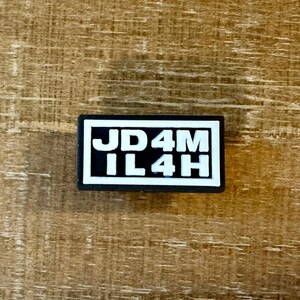 Könnte beinhalten: Ein rechteckiger schwarz-weißer Anhänger mit dem Text "JD4M IL4H" in weißen Blockbuchstaben. Der Anhänger befindet sich auf einem Holzgrund.