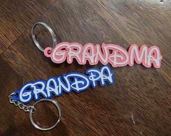 May include: Two keychains with the words "Grandma" and "Grandpa" in a playful font. The "Grandma" keychain is pink with white accents, while the "Grandpa" keychain is blue with white accents. Both have silver key rings.