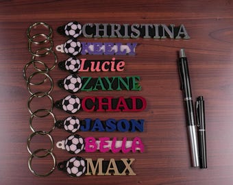 May include: Eight custom keychains featuring a soccer ball charm and a name tag with the names CHRISTINA, KEELY, Lucie, ZAYNE, CHAD, JASON, BELLA, and MAX. Each keychain includes a metal ring, and two black pens are positioned to the right.