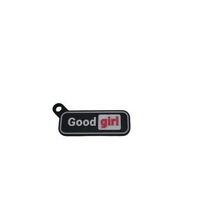 May include: A black rectangular charm with the words "Good girl" in white and red lettering. The charm has a small loop at the top for attachment.