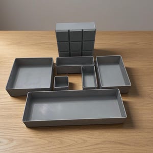 May include: A selection of grey storage containers in different shapes and sizes. The set includes rectangular trays, square boxes, and a multi-compartment organiser. The containers are arranged on a light wood surface, offering a minimalist aesthetic.