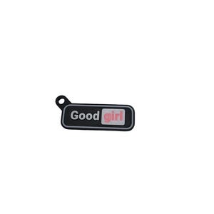 May include: A black shoe charm with the words "Good girl" in white and pink. The charm has a small loop at the top for attachment. The text is centered on a rectangular shape.