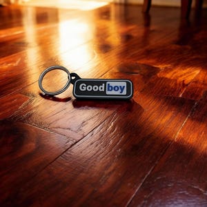 May include: A black and white keychain with the words "Good Boy" in white text on a black rectangle with a blue accent. The keychain is on a dark wood floor.