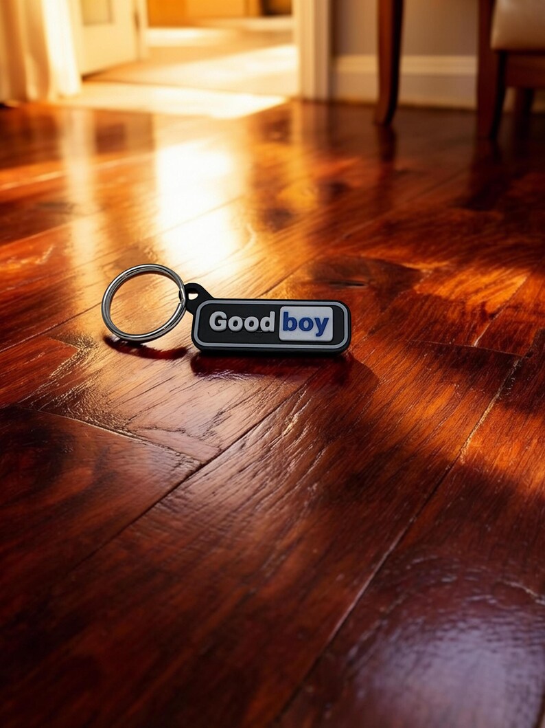 May include: A black and white keychain with the words "Good Boy" in white text on a black rectangle with a blue accent. The keychain is on a dark wood floor.