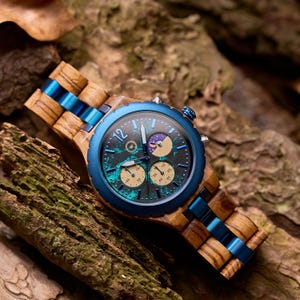 Può includere: Un orologio da polso in legno con una lunetta blu e un cinturino blu abbinato. Il quadrante dell'orologio è di colore verde-blu scuro con quadranti secondari color oro. L'orologio è ambientato su uno sfondo di legno e foglie.