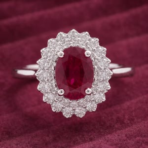 Può includere: Un anello color argento con una gemma centrale rosso scuro a taglio ovale, circondata da un doppio alone di piccole pietre trasparenti a taglio rotondo. La montatura è a griffe e l'anello presenta una finitura in metallo liscio e lucido.