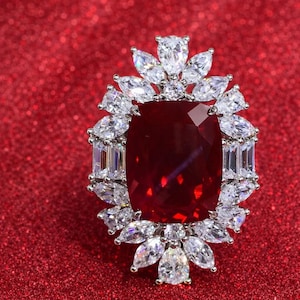 May include: A striking piece of jewellery with a large, rectangular, deep red gemstone at its centre. It's encircled by smaller, clear, sparkling stones, set against a vibrant red background, creating a luxurious appearance.