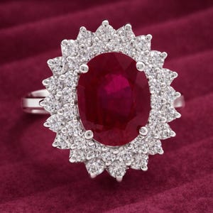 Może przedstawiać: Srebrny pierścionek z dużym owalnym rubinem otoczonym aureolą mniejszych, okrągłych, białych diamentów. Pierścionek osadzony jest na ciemnoczerwonym tle, podkreślającym żywe kolory kamieni szlachetnych.