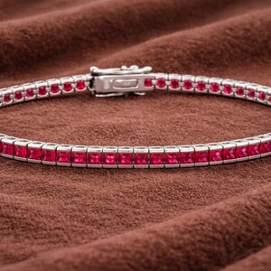 May include: A silver bracelet with a row of square-cut, vibrant red gemstones. The bracelet is set against a brown, textured background, highlighting the jewelry's sparkle and design. The clasp is visible, indicating a secure closure.