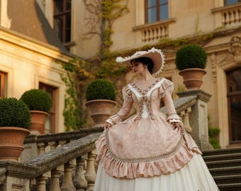 Könnte beinhalten: Eine Person in einem rosa-weißen viktorianischen Kleid und Hut steht auf einer Steintreppe. Das Kleid hat Spitze, Rüschen und goldene Stickereien. Im Hintergrund sind ein großes Gebäude und Topfpflanzen zu sehen.