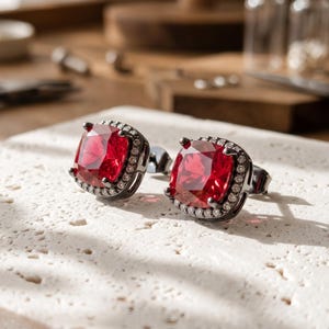 May include: A pair of stud earrings featuring square-cut, vibrant red gemstones set in dark metal frames. Each central red stone is surrounded by a halo of small, clear white accent stones, creating a contrast against the dark setting.