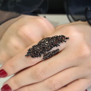 May include: A black statement ring featuring a cluster of small, dark gemstones. The ring has an elongated design that covers a portion of the finger and a smaller band. The gemstones are set in a dark metal.