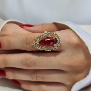 May include: A gold ring with a large, teardrop-shaped, deep red gemstone. The gemstone is encircled by a double halo of small, clear crystals. The ring is on a hand with red nail polish.