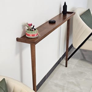 Puede incluir: Una consola estrecha de madera oscura con una pequeña estantería. Una pequeña planta en maceta con detalles en rosa y verde se encuentra en el lado izquierdo. Una botella negra y un pequeño disco negro están en la estantería. La mesa tiene cuatro patas delgadas.