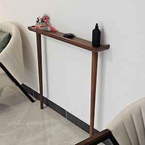 May include: A narrow, dark wood console table with a rectangular top and four slender legs. A black bottle and a small decorative item sit on the table. Two white armchairs with black frames are in the background.