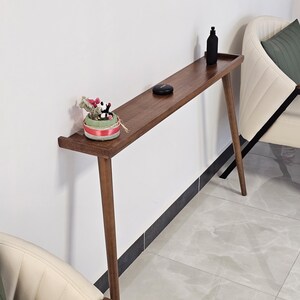 Puede incluir: Una consola de madera de estilo moderno de mediados de siglo, con un acabado oscuro. La mesa tiene una parte superior larga y rectangular y patas anguladas. Una pequeña planta decorativa, una botella negra y un pequeño disco negro están sobre la mesa.