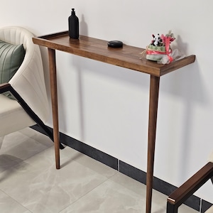 Op de afbeelding: Een smalle, houten console met een donkere afwerking, ondersteund door vier slanke, taps toelopende poten. Een zwarte fles en een kleine decoratieve opstelling staan op het tafelblad. Twee crèmekleurige stoelen met zwarte accenten staan aan weerszijden.