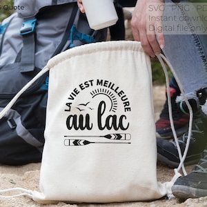 May include: A cream-coloured drawstring bag with the phrase "LA VIE EST MEILLEURE AU LAC" in black, along with a sun, birds, and oars. A backpack is visible in the background. The bag is a digital file download.