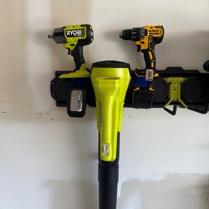 May include: A wall-mounted black tool organizer rack holding a lime green Ryobi impact wrench with a battery, a yellow and black DeWalt drill with a battery, and a large lime green Ryobi leaf blower with a black nozzle attachment.