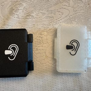 May include: Two rectangular cases for earplugs, one in solid black and one in translucent white. Each case features a hinged side and a printed graphic on the front showing a stylized ear with an earplug inserted, indicating the contents inside.