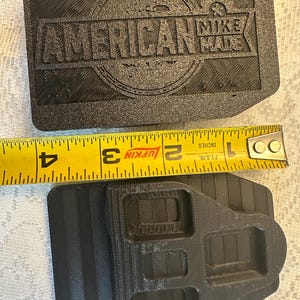 May include: Black rectangular object with the text "AMERICAN MIKE MADE" and a second black object with a complex shape. A yellow tape measure shows the objects are approximately 3 inches wide.