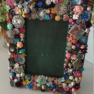 May include: A green picture frame decorated with colorful jewels, buttons, and other trinkets. The frame is surrounded by a variety of embellishments, including flowers, hearts, and a clock face.