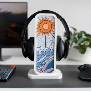 以下が含まれることがあります： 太陽、雲、波のカラフルなイラストが描かれた長方形のデザインの白いヘッドホンスタンド。 黒いヘッドホンはスタンドに置かれ、キーボードとマウスのある机の上に置かれています。