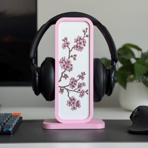 以下が含まれることがあります： 桜の花のデザインが施された白い中央パネルを備えたピンク色のヘッドフォンスタンド。黒いヘッドフォンがスタンドに置かれています。スタンドは、キーボードとマウスを備えた机の上にあります。