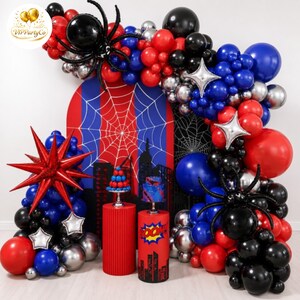 May include: A superhero-themed party balloon arch featuring red, blue, black, and silver balloons, two large black spider balloons, a red starburst balloon, and silver star balloons. Two cylindrical pedestals display a cake and a balloon tower.