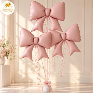 May include: Three large, light pink, bow-shaped foil balloons arranged in a cluster. Each balloon features a central knot and two trailing ribbon-like ends. The balloons are attached to thin, curling pink ribbons that gather at a weighted base.