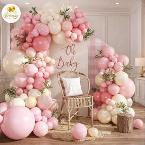 Puede incluir: Fondo de fiesta de bienvenida al bebé con un arco de globos rosas y blancos y vegetación. Una silla de mimbre con un cojín blanco está frente a un letrero que dice "Oh Baby". La escena está decorada con globos adicionales y un tocón de madera.