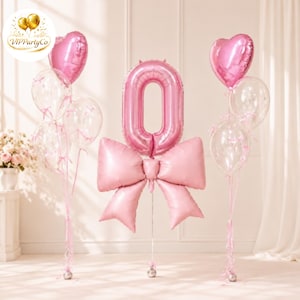 May include: A pink foil balloon shaped like the number zero, accented with a large pink foil bow at its base. It is flanked by two clusters of balloons, each featuring a pink heart-shaped foil balloon and two clear balloons with pink ribbons.