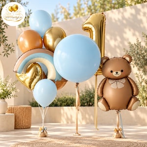 May include: A balloon arrangement featuring a large light blue balloon, a gold number one balloon, and a brown teddy bear balloon. Includes a rainbow-shaped balloon, gold heart balloon, and additional light blue and gold round balloons.