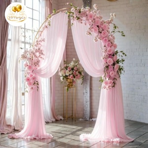 Puede incluir: Un arco circular de metal dorado drapeado con tela rosa claro. El arco está decorado con racimos de rosas rosas y pequeñas flores blancas. Un arreglo floral a juego se encuentra sobre un soporte de metal dorado detrás del arco.