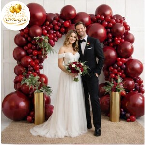 May include: A wedding scene with a burgundy balloon arch framing a couple. The bride wears a white off-the-shoulder gown and holds a bouquet. The groom is in a black suit. Gold vases with floral arrangements are on either side of the couple.
