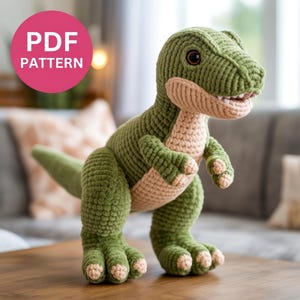 Puede incluir: Un juguete de dinosaurio tejido a ganchillo en verde y beige, con una expresión amigable. El dinosaurio tiene un aspecto texturizado y afelpado, con puntadas visibles y detalles cuidados. La imagen incluye un círculo rosa con el texto "PDF PATTERN".